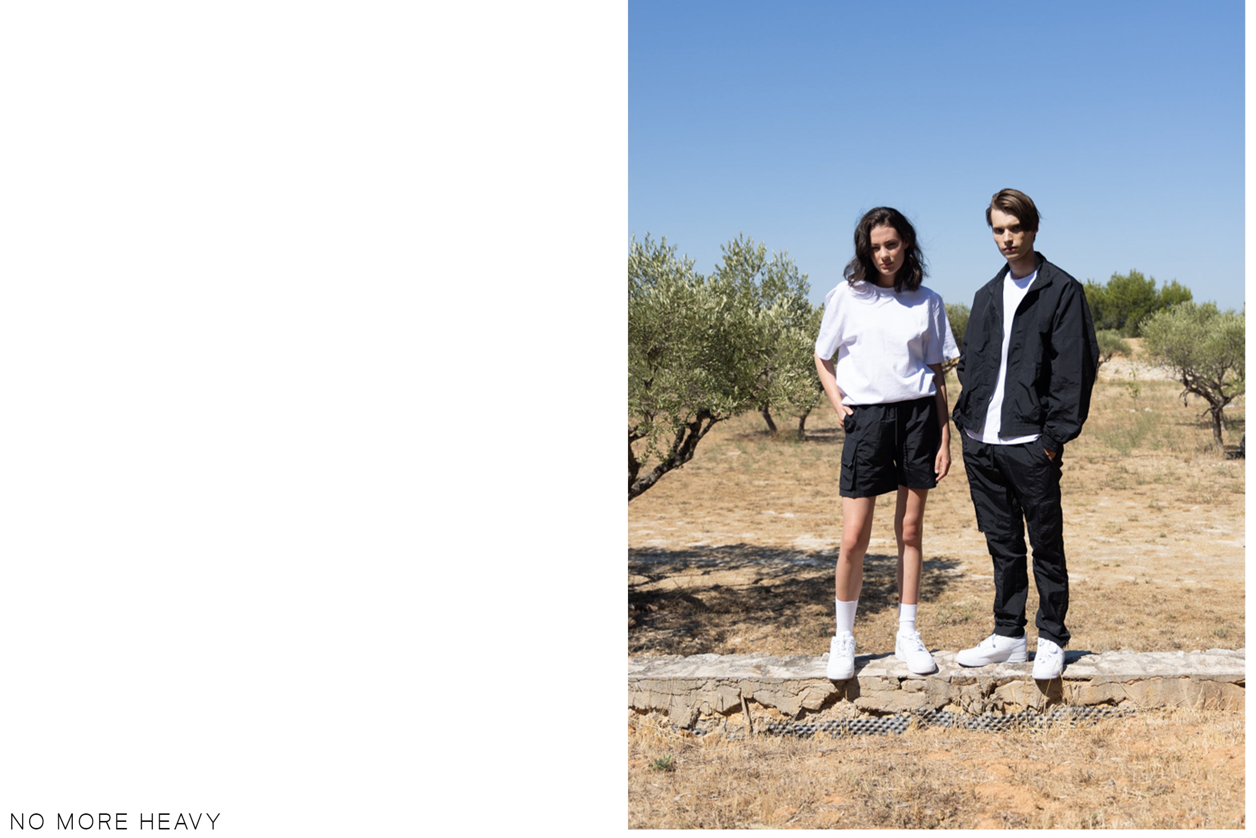 A male model wearing a black zip-up jacket and black cargo pants stands beside a female model wearing a white t-shirt and black cargo shorts, posing in a grassy field.