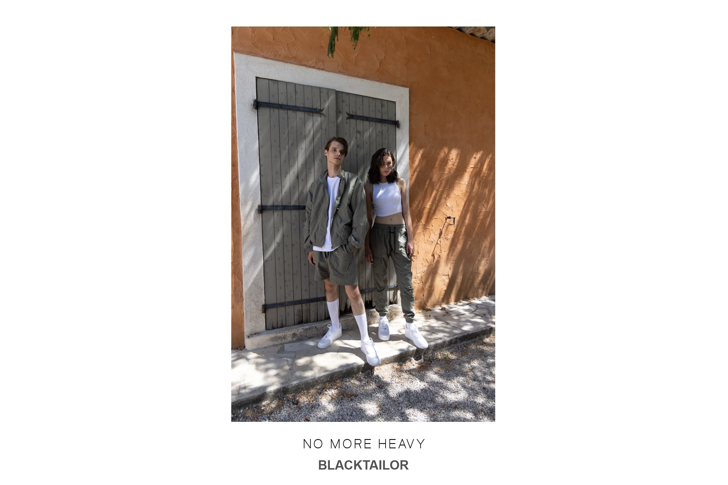 A  male model wearing a gray zip-up jacket and gray cargo shorts stands next to a female model wearing a white tank top and gray cargo pants, posing in front of a stone wall.