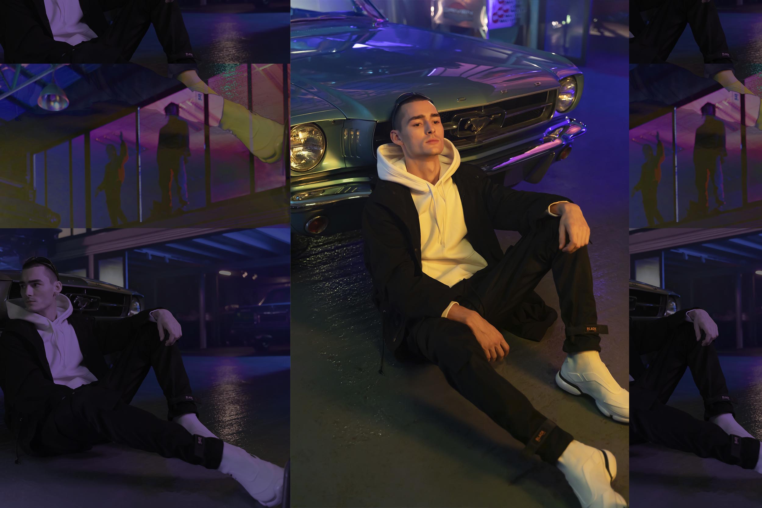 A female model in black jacket and black cargo pants is sitting on the floor next to a car.