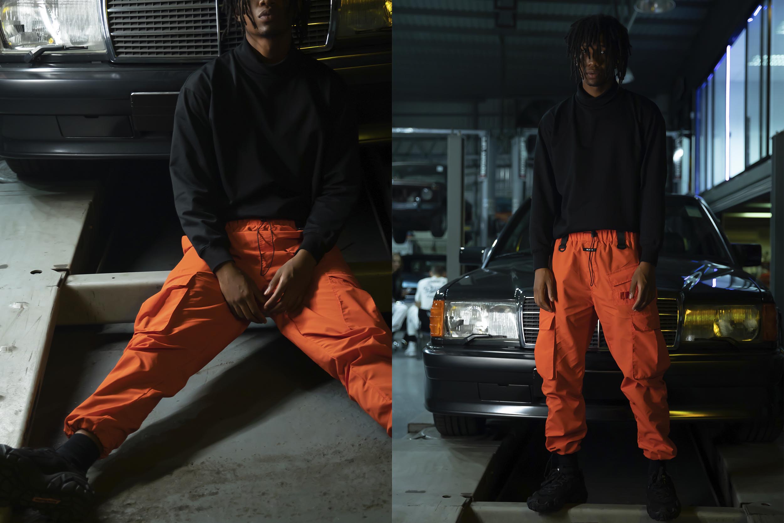 A male model in orange cargo pants and black jacket is standing next to a car.