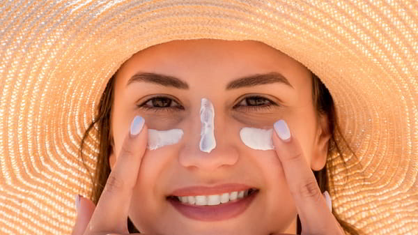 Girl wearing a large sun hats points her fingers at white sunscreen on her cheeks and nose