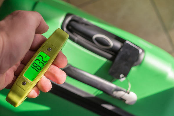 Someone holding a luggage scale, one of the best travel basket gift ideas, above a green suitcase