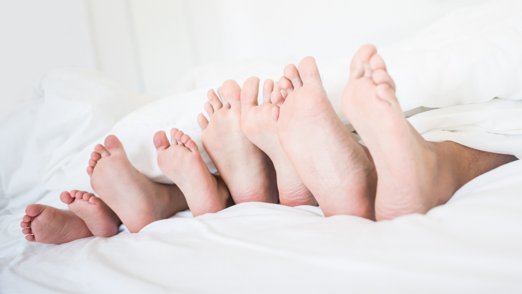 Set of 4 feet, a family, snuggled up on bed with just their feet popping out of the bedsheets.