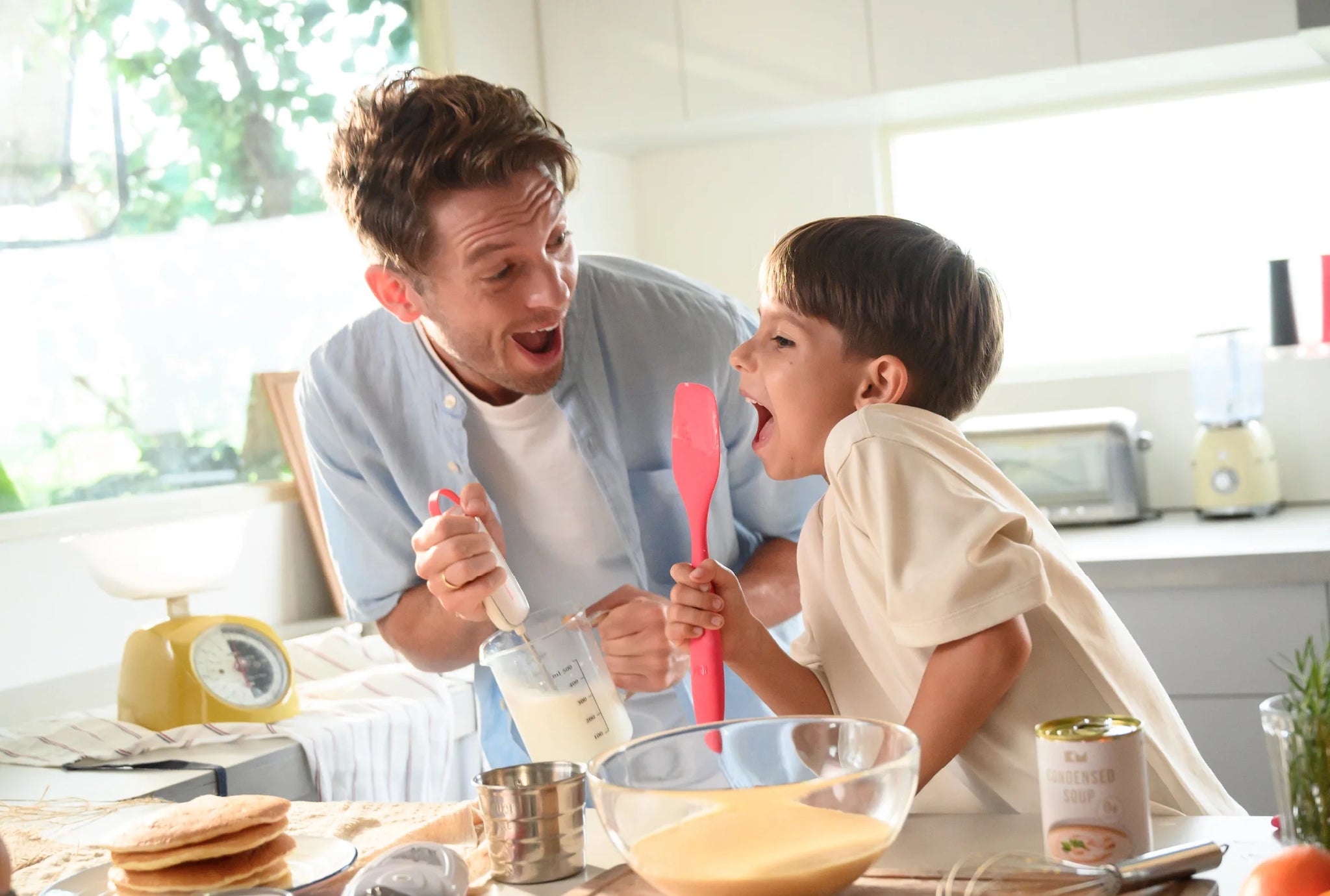 A father and young son laughing together in the kitchen while baking, with the boy holding a red Kitchen Mama spatula and the dad using a milk frother near a bowl of ingredients.