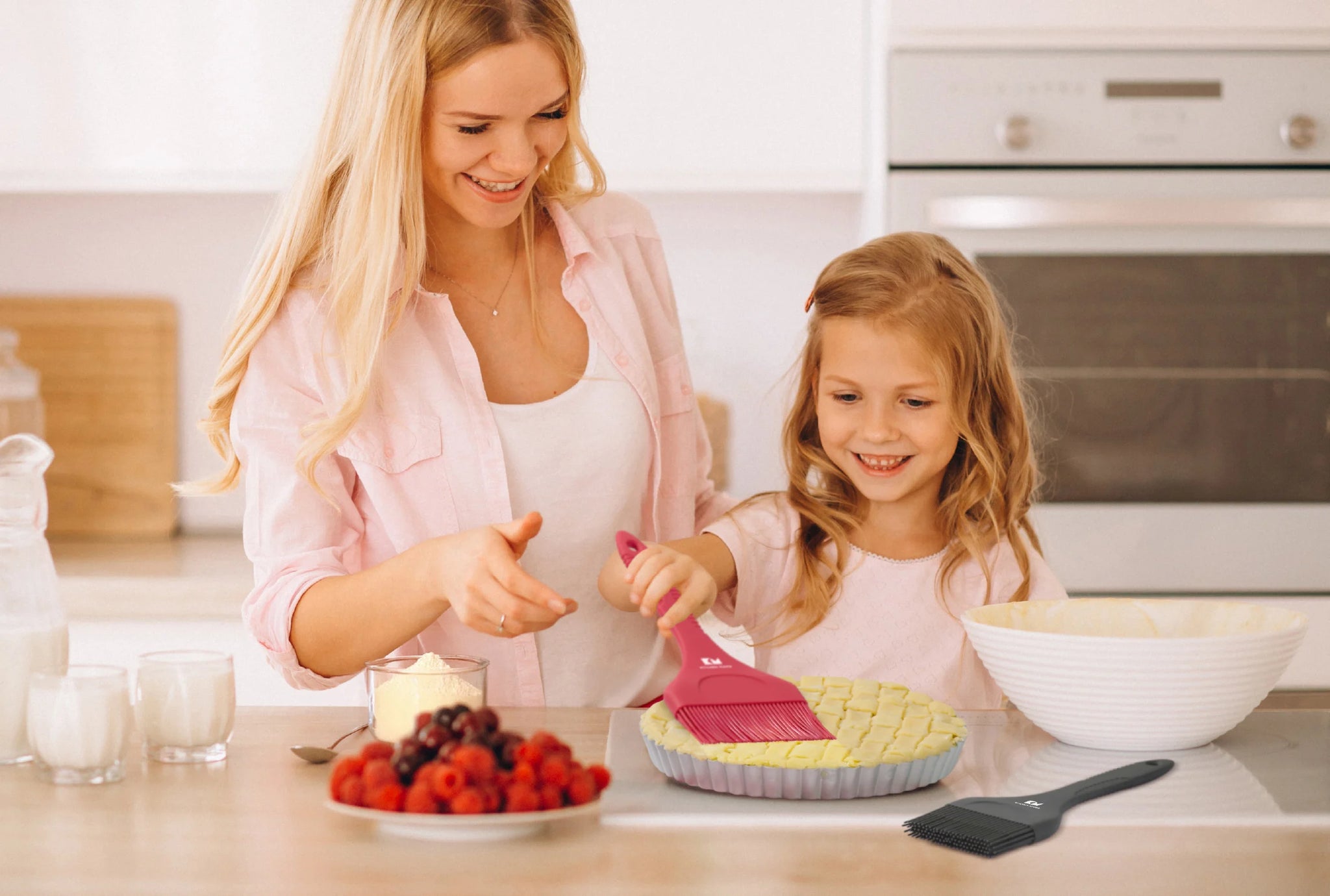 A smiling mother and daughter baking together in a bright kitchen, with the child brushing a pie using a pink Kitchen Mama silicone brush.