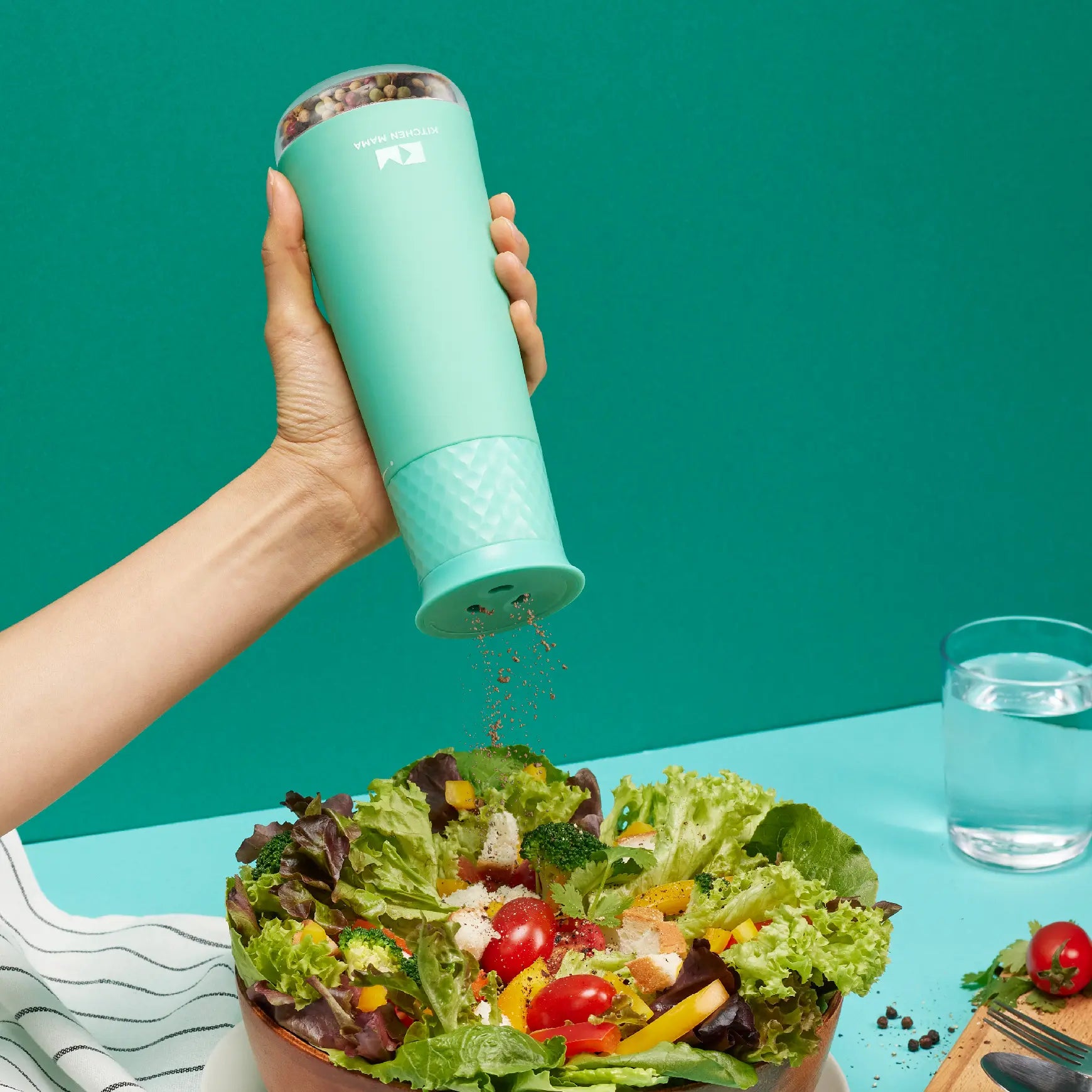 A hand holds a mint-green spice shaker above a colorful salad bowl, sprinkling seasoning onto fresh greens and vegetables.