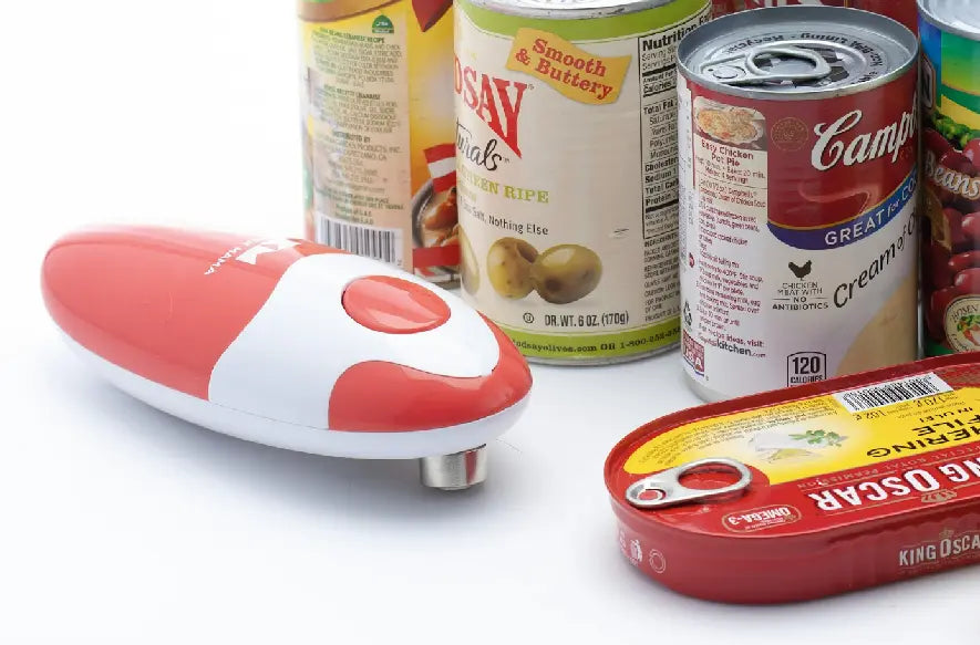 Red and white Kitchen Mama can opener next to canned goods like Campbell’s soup and olives on a white surface.
