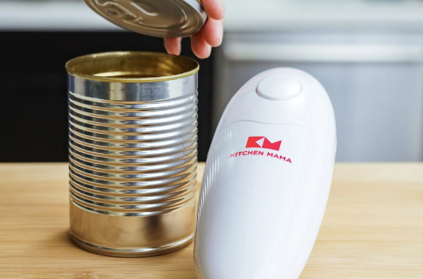 A white Kitchen Mama electric can opener next to an open tin can with a hand lifting the lid on a wooden kitchen counter