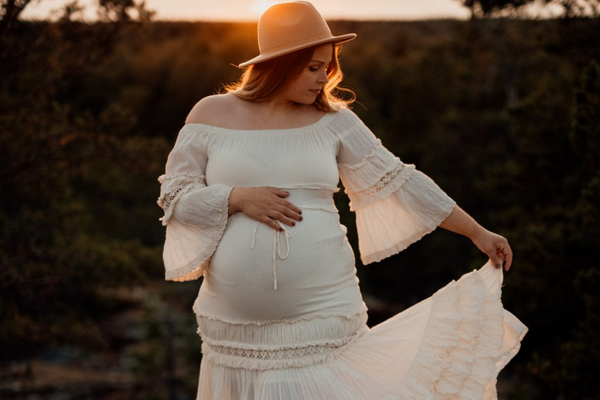 A pregnant woman during sunset wearing a white flowy dress and a hat
