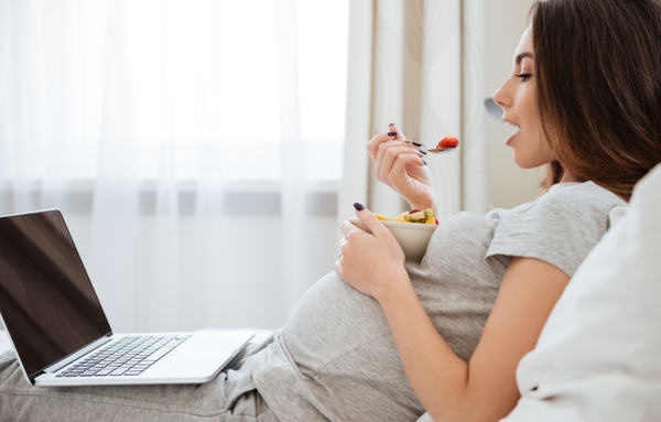 Woman is chilling in bed with her laptop and eating healthy foods during pregnancy