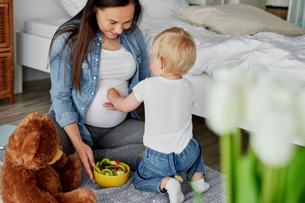 A pregnant woman is sitting on the floor with her child while holding a bowl of fruit