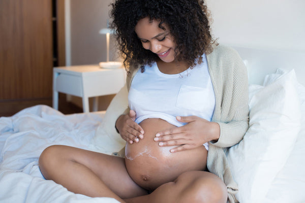 A pregnant woman is sitting on a bed and spreading moisturizer on her stomach