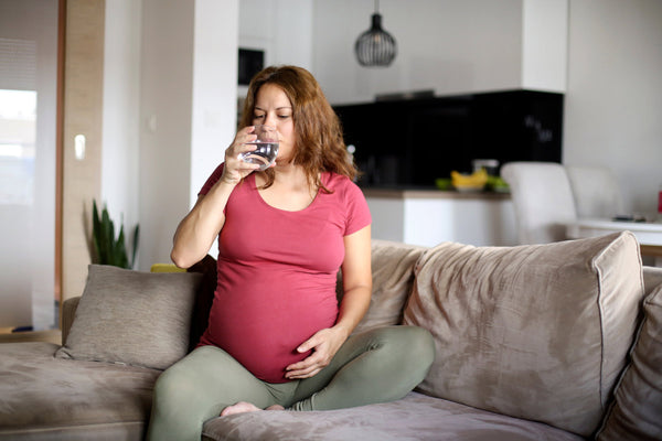 A pregnant woman with existing siblings is sitting on a couch and drinking a glass of water