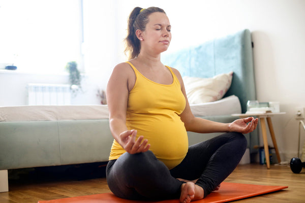 A pregnant woman is meditating on a floor, wearing a yellow top