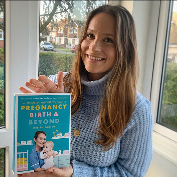 Kätilö Marie Louise poseeraa sinisen kirjansa “Pregnancy, birth & beyond” kanssa ikkunan edessä