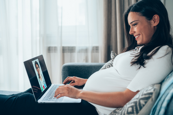 A pregnant woman is smiling and taking a remote call with a doctor