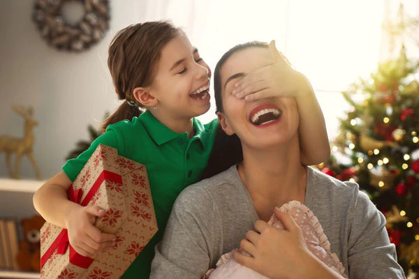 Tytär peittää nauravasti äitinsä silmät ja pitelee joululahjaa toisessa kädessään jouluna
