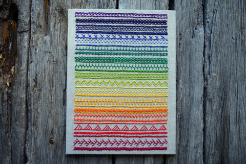 Finished band sampler in rainbow colors on oatmeal linen, simply mounted and set against wood backround