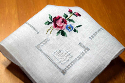 Handkerchief folded into quarters showing drawn threadwork in soft blue and rose in pink and green with metallic accents cross stitched above