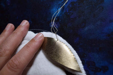 Hand stitching an almond-shaped piece of gold kid leather to white ear of rabbit. Background of dark blue painted silk.