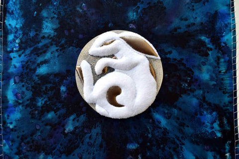 A rabbit is made of sculpted white felt with gold kid leather accents. It is curled on its side, facing left. Around the rabbit and completing a circle are silver threads secured with blue silk so the rabbit appears to be sitting on a coin. The background is dark blue painted silk. 'Moon Rabbit'