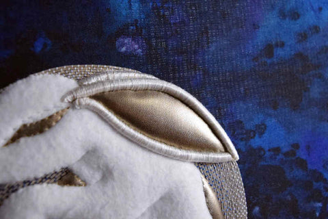 Closeup shot of white felt rabbit in silver circle on dark blue background; inside of ear and other details are in gold leather and edge of ear is raised and padded.