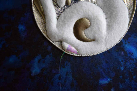 Closeup of bottom half of moon rabbit in raised white felt and gold leather against silver disc on blue background; the tail is being stitched with iridescent pink flat ribbon