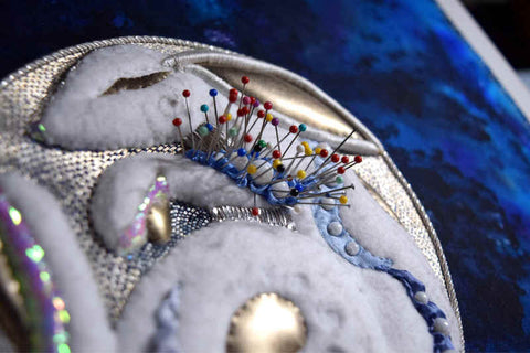 Closeup of Moon Rabbit stitched in raised white felt with gold accents on silver disc against blue background. Dozens of pins hold ribbon and beads on arm of rabbit.