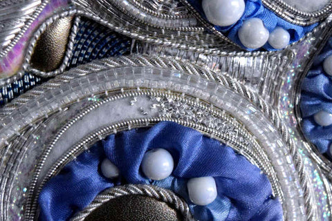 Closeup of silver and blue embroidery (goldwork) with tiny chips of bright silver filling in a crescent area over blue silk and beads.