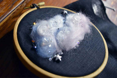 A small embroidery hoop holding black fabric; a fluffy cloud is worked on it in pink and blue wool with stars and tiny beads scattered through it