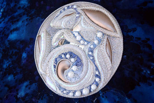 Intricate silver embroidery with beads on deep blue background, showing a stylized rabbit in relief over the face of the full moon. 