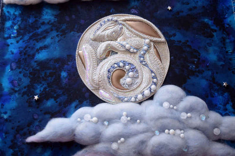 A stylized, Art Noveau representation of the moon behind clouds; the moon is intricately worked in silver, silk, and pearly beads to show a sleeping rabbit curled up, and the clouds are made of puffy wool with scattered pearly beads catching the light.