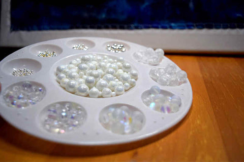 A small artist's palette with each indentation filled with beads sits on a table; the beads are pearly, clear, and silver in small piles