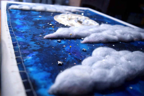 Extreme angle shot of moon and clouds embroidery; moon is gleaming silver with the suggestion of a rabbit worked on its face, clouds are puffy wool. Around them scattered on the dark blue background are tiny silver chips, crystals, and some larger stars pinned in place.
