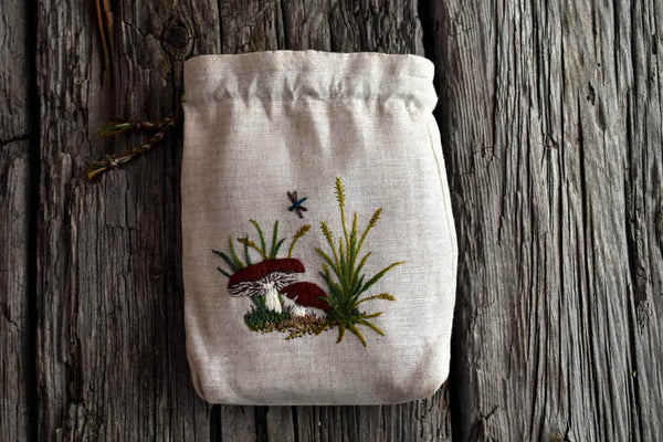Small linen pouch hand embroidered with a design of mushrooms, grass, and small dragonfly. Pouch has a braided cord drawstring.