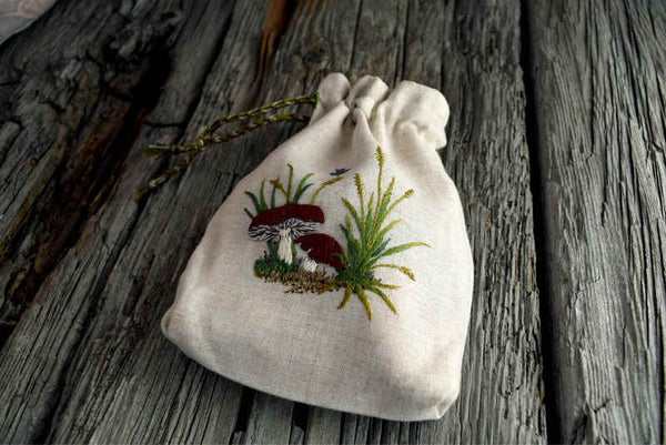 Drawstring pouch with braided cord shown closed; pouch is made of oatmeal colored linen and ornamented with hand embroidered mushrooms and grass