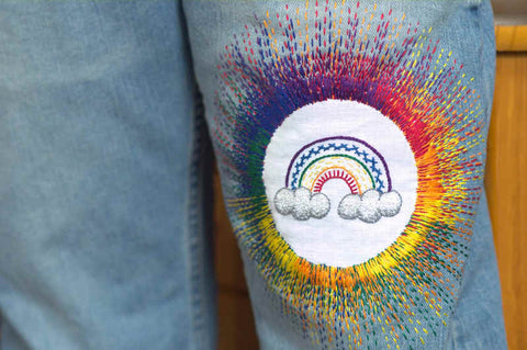 Hand embroidered patch on pair of jeans, with rainbow worked in bright colors and various stitches surrounded by a rainbow starburst of running stitch worked from the patch into the denim