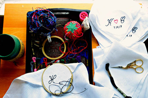 White linen handkerchiefs being hand embroidered; one in progress sits on a  tray with thread, pincusion, and teacup of snips while others lay to the side