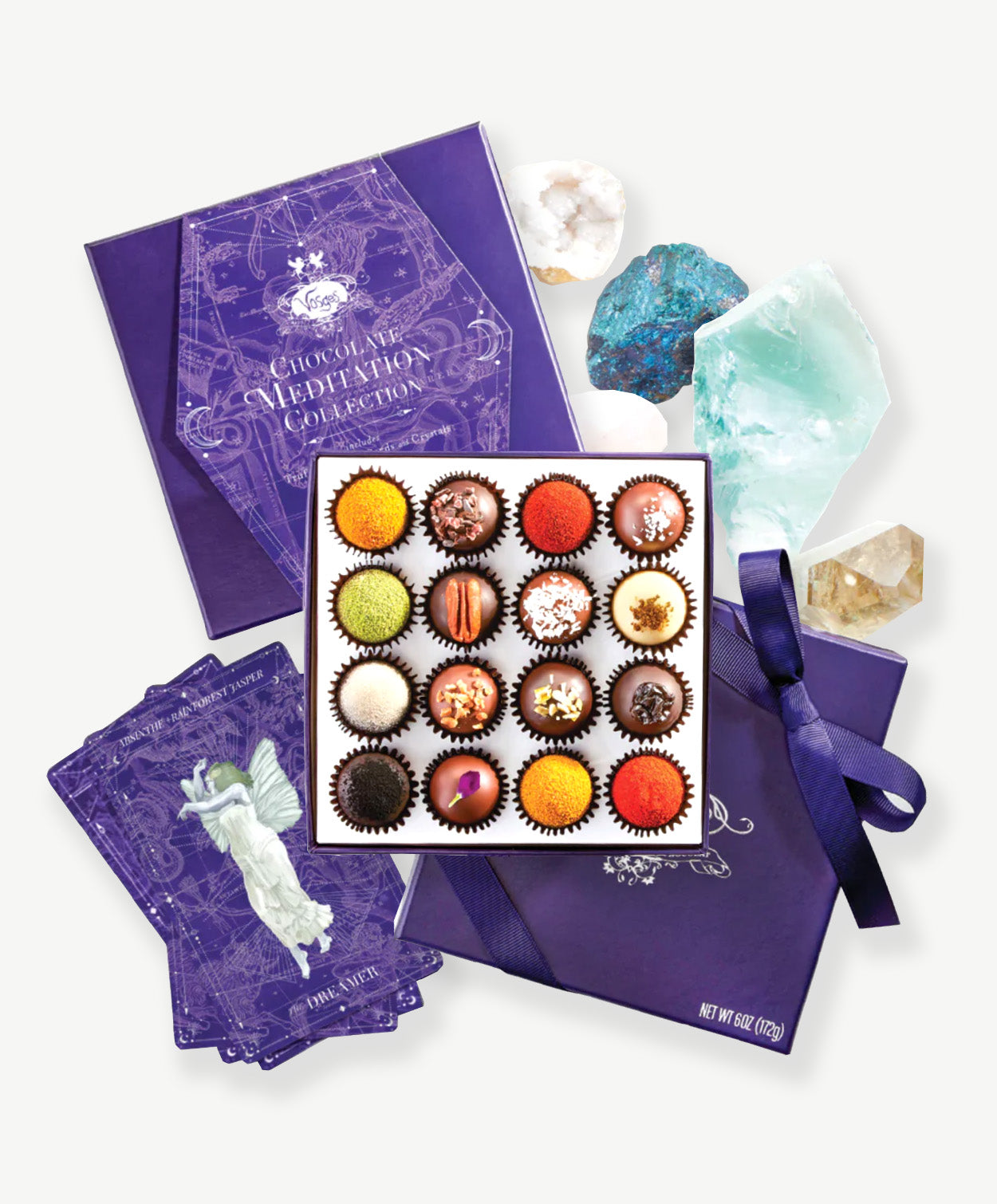 An open box of Vosges chocolate truffles adorned in brightly colored spices, nuts and dried flowers sits surrounded by crystals, precious stones, affirmation cards and meditation guide sit on a grey background.