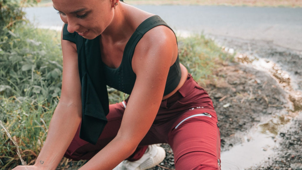 A white woman, sitting in front of a natural spring of water. She is wearing a red Hiking Pants from BARA Sportswear.