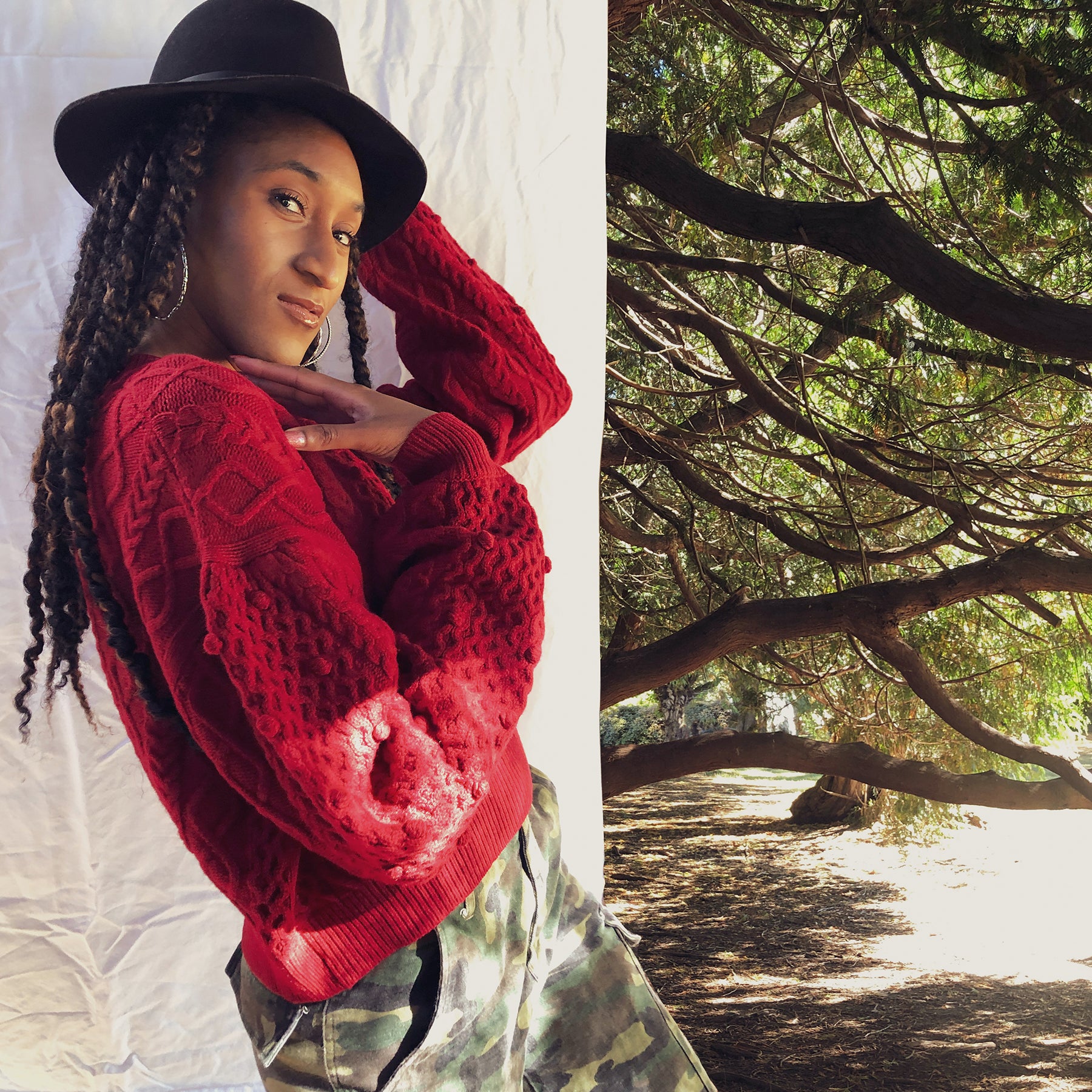 Dark skin model wearing silver hoops, black hat and red sweater standing under trees.