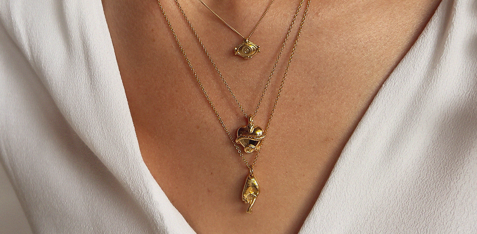 Fall Jewelry for women. Three gold charm necklaces on a model wearing a white shirt. The charms are fingers crossed for good luck, and eye for protection, and a snake wrapped around a heart for wisdom.