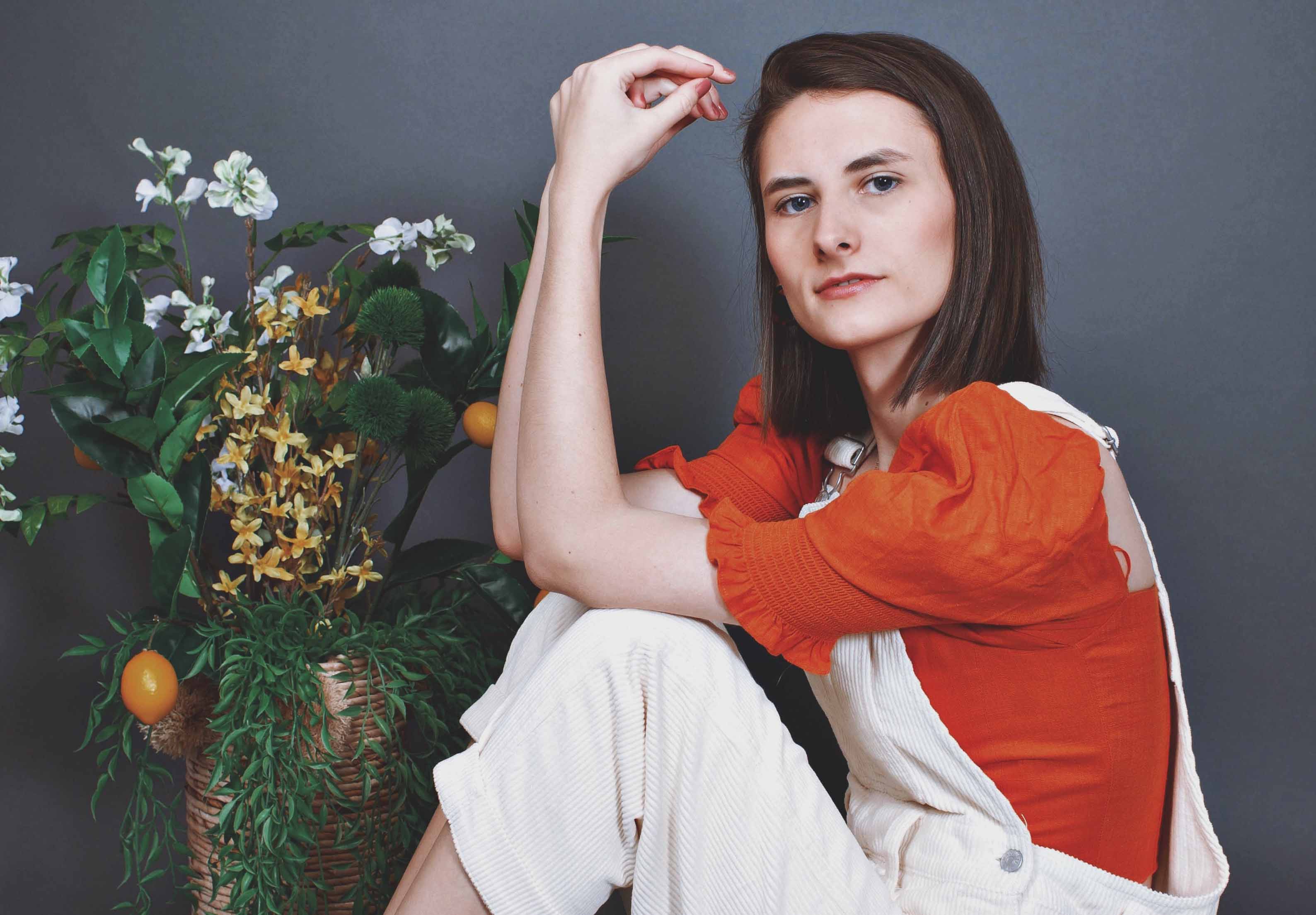 Making Sustainability Sustainable. Ethical fashion blogger Hanna Cody sitting on blue backdrop wearing white overalls and orange shirt