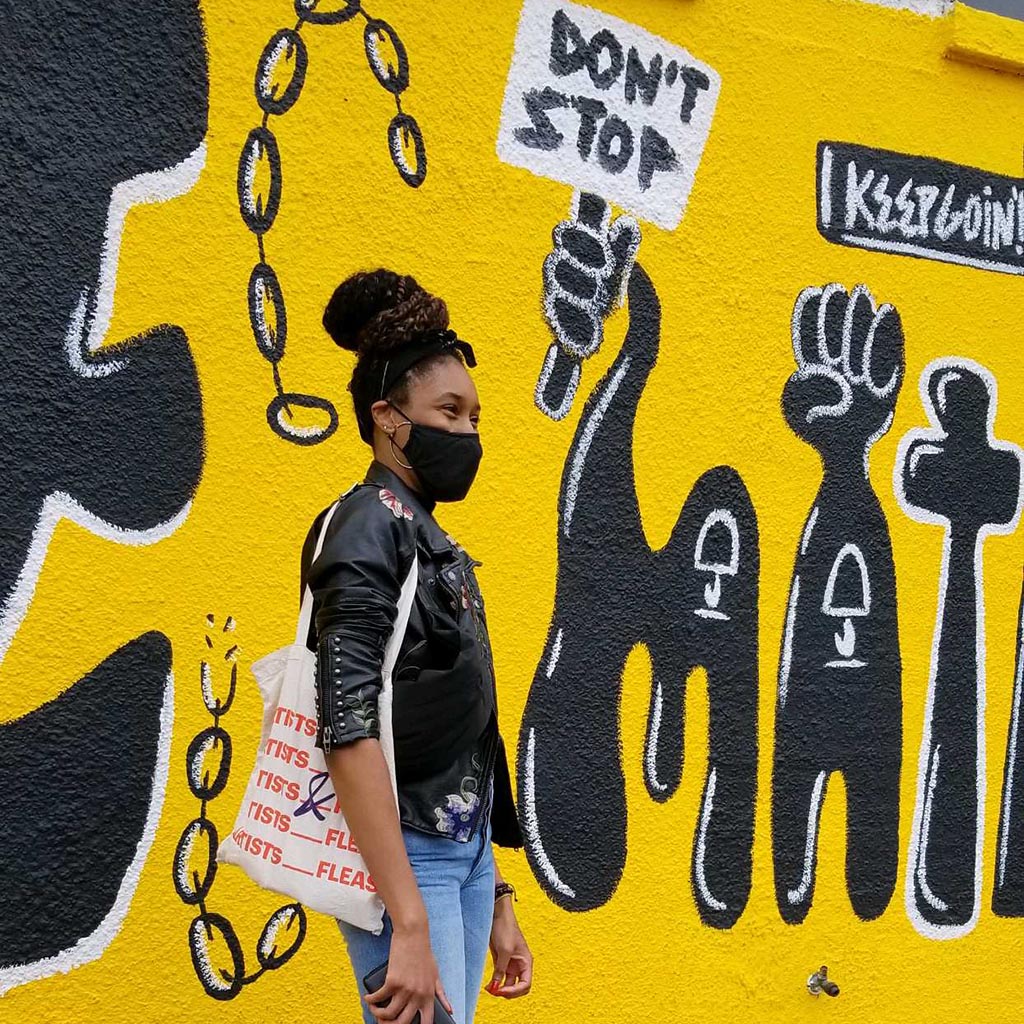 Podcast Host on Fashion standing in front of a bright yellow and black mural with the words "don't stop" and "keep going"