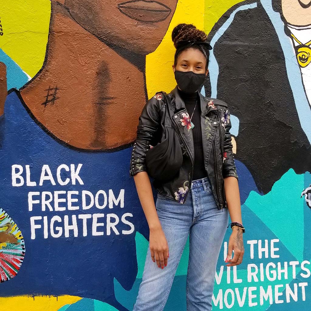 Black Podcast host in front of mural about the Civil Rights Movement. It features colorful art of black historic figures and has the words, "Black freedom fighters" and "the civil rights movement"