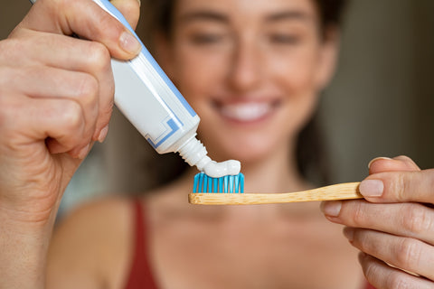 A woman holds up her toothbrush in front of the camera with her left hand while her right hand is squeezing toothpaste onto it.