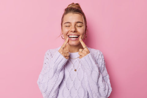 A woman wearing a purple sweater smiles with her eyes closed while pointing at her teeth with both hands.