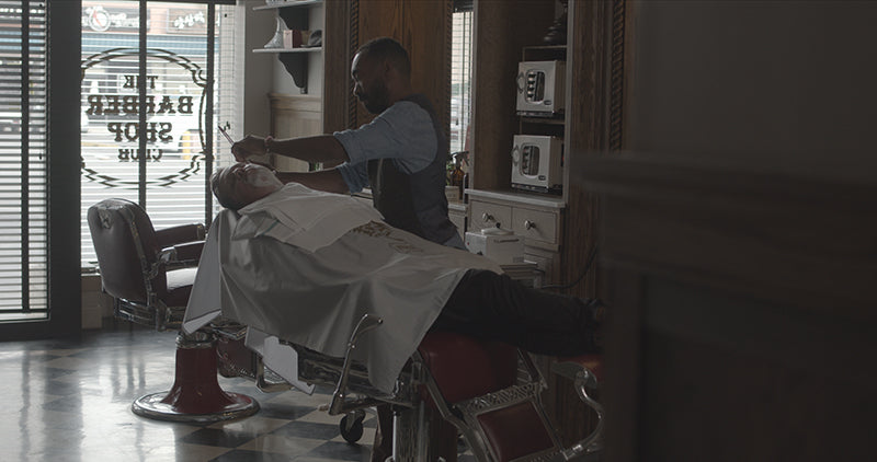 Barber Woody shaving Ian who is reclined in a barber chair with shaving cream on his face