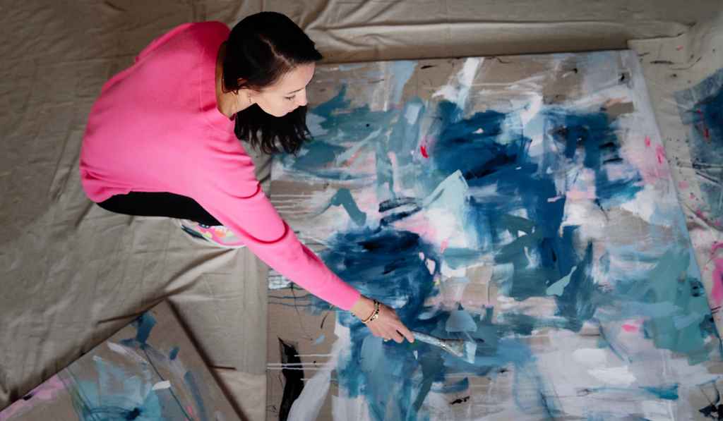 A woman wearing a pink shirt paints a blue abstract painting on the ground.