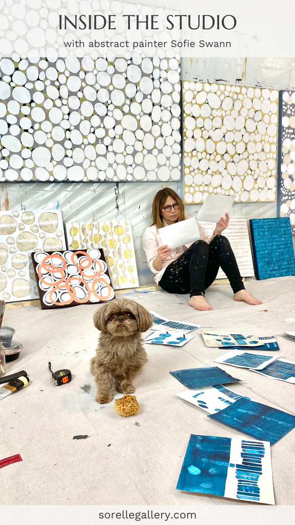 An artist sits in an art studio with a dog surrounded by abstract paintings. 